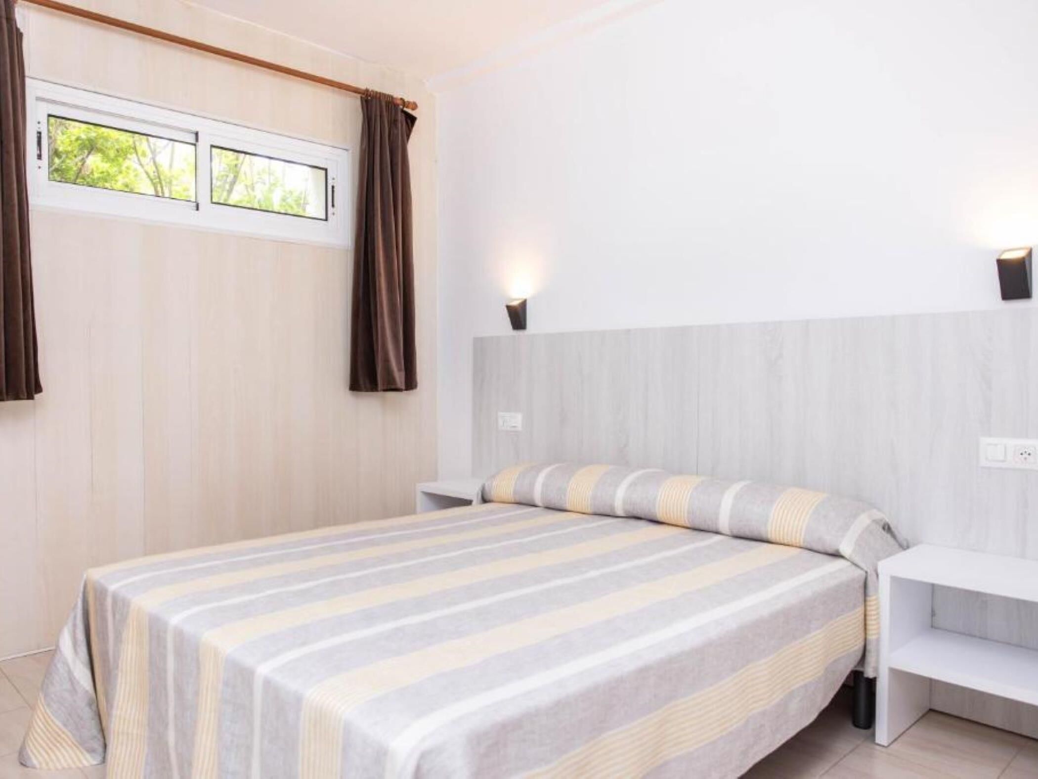 A modern bedroom with a large bed, two bedside tables, a nightstand, a window with curtains, and wall-mounted lighting.