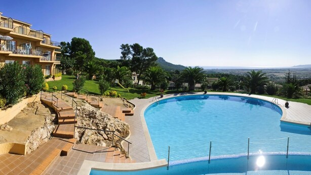 A large, curved swimming pool with a clear view of the surrounding landscape, including a building with balconies and a distant mountain range. - APCOSTAS Club Torre Vella / Three Bedrooms (L'Estartit)