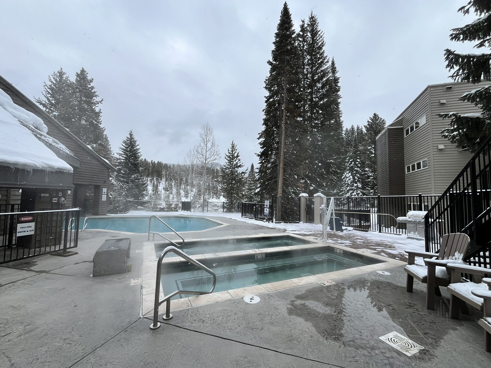 Uma área de piscina com uma escada de metal que dá acesso à água, rodeada por terreno e árvores cobertas de neve.