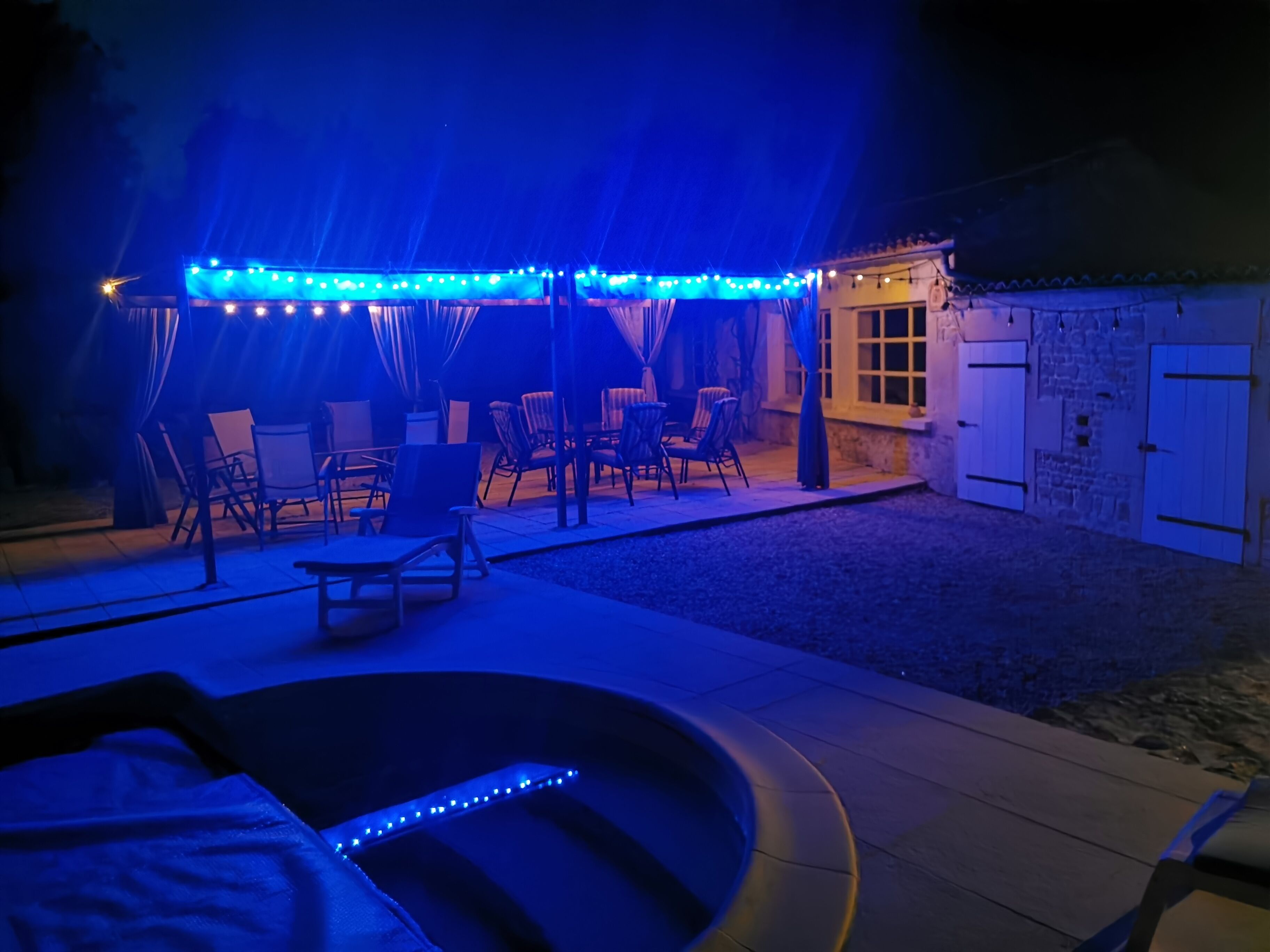 Een buitenpatio met een zwembad, stoelen en een overdekt zitgedeelte met lichtslingers.