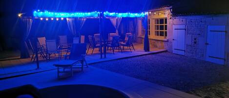 Een buitenpatio met een zwembad, stoelen en een overdekt zitgedeelte met lichtslingers.