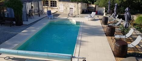 En rektangulär swimmingpool med klart, blått vatten, omgiven av en kakelbeklädd poolterrass. På höger sida finns solstolar och parasoller, och på vänster sida finns en överbyggd sittgrupp med stolar.