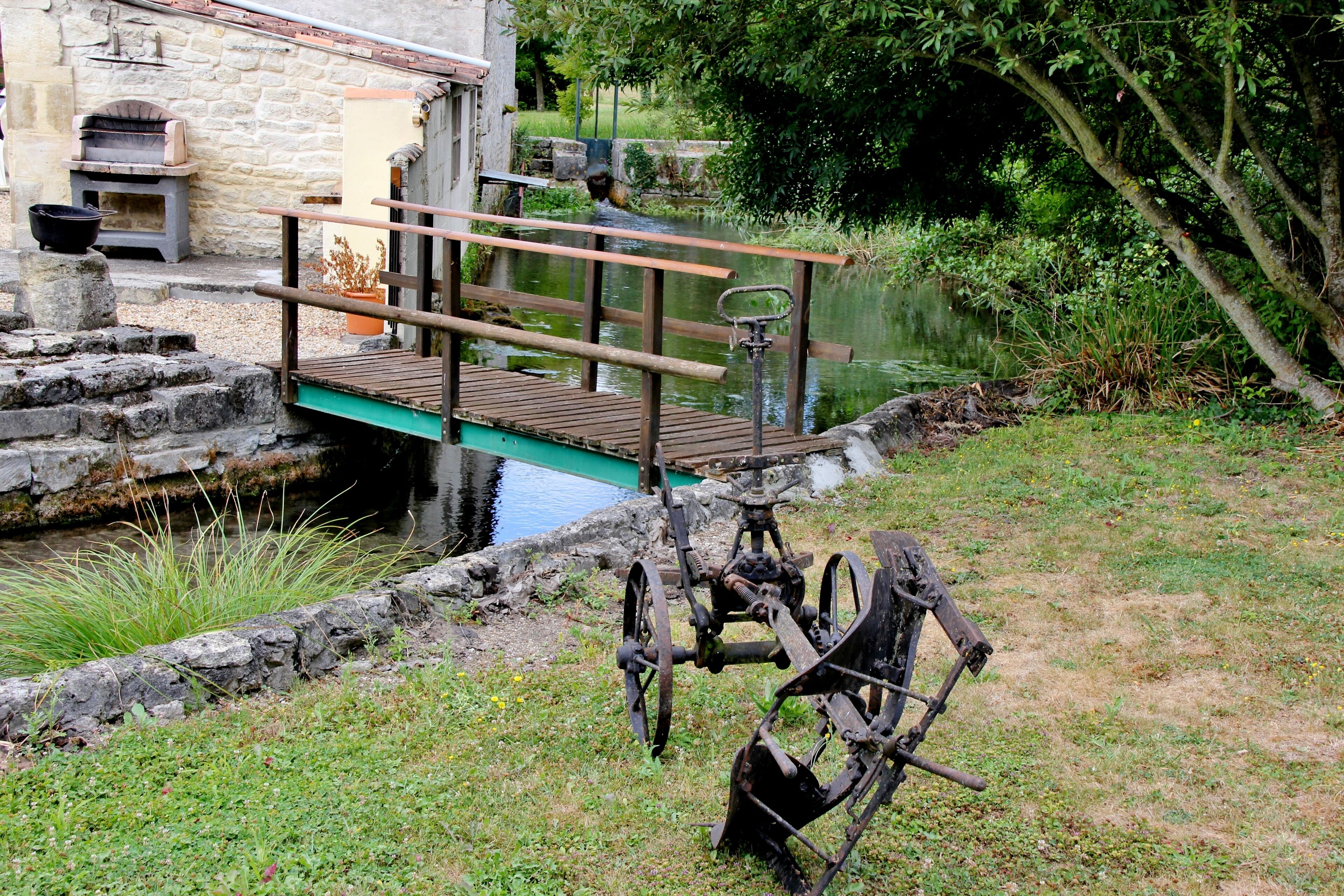 A rustic wooden bridge over a calm river, a stone building with a chimney, and a grassy area with a large metal structure.
