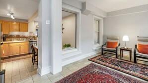 A modern kitchen with wooden cabinets, a dining area with two chairs and a rug, and a living room with a lamp and chair.