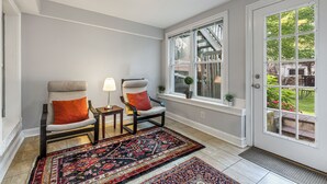 A room with two armchairs, a patterned rug, a small table with a lamp, and a window with a view of a staircase and greenery.