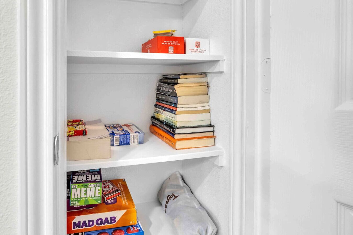 Een witte plank met een stapel boeken, een doos met de tekst 'The Game of Life' en een zak 'Mad Gab'.