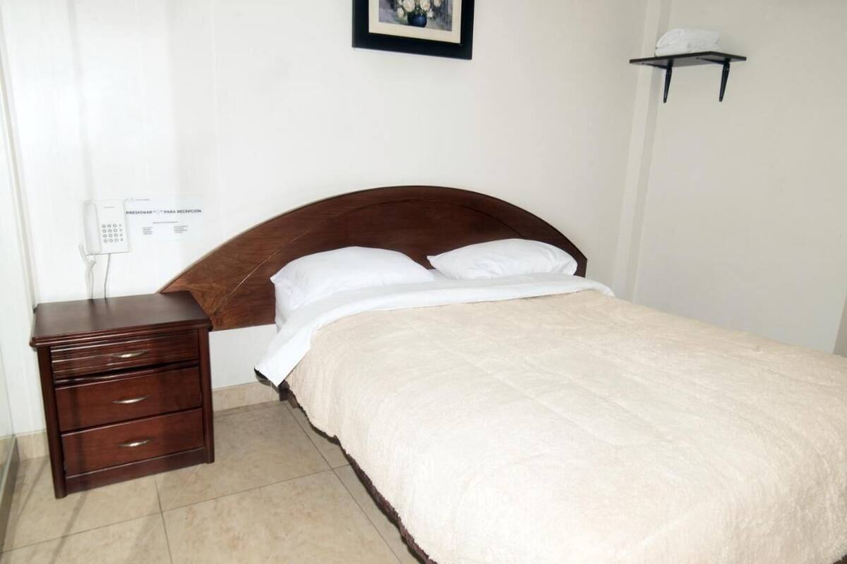 A bedroom with a wooden bed, white bedding, a bedside table with drawers, a wall-mounted phone, and a small shelf with a towel.