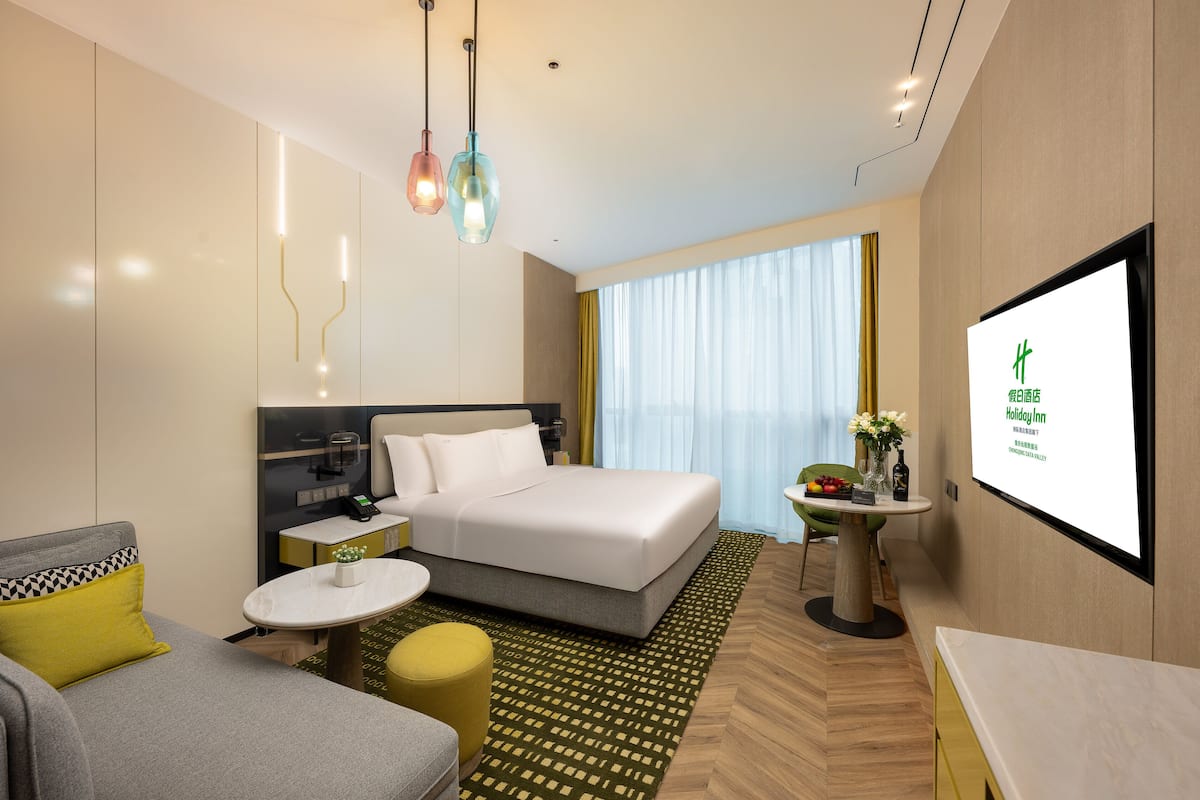 A modern hotel room with a bed, a desk, a chair, a small table, and a TV screen displaying the hotel name.
