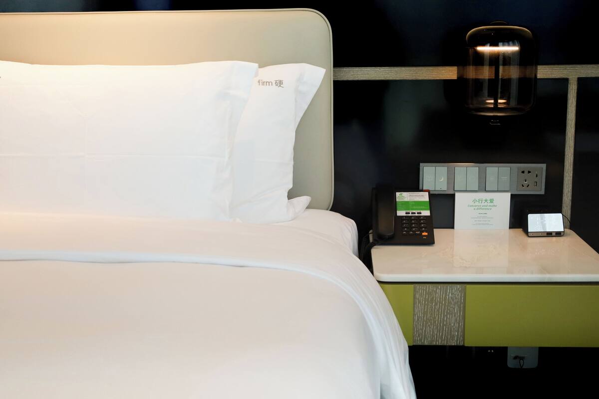 A hotel room with a neatly made bed, a bedside table with a phone and a small appliance, and a wall-mounted light fixture.