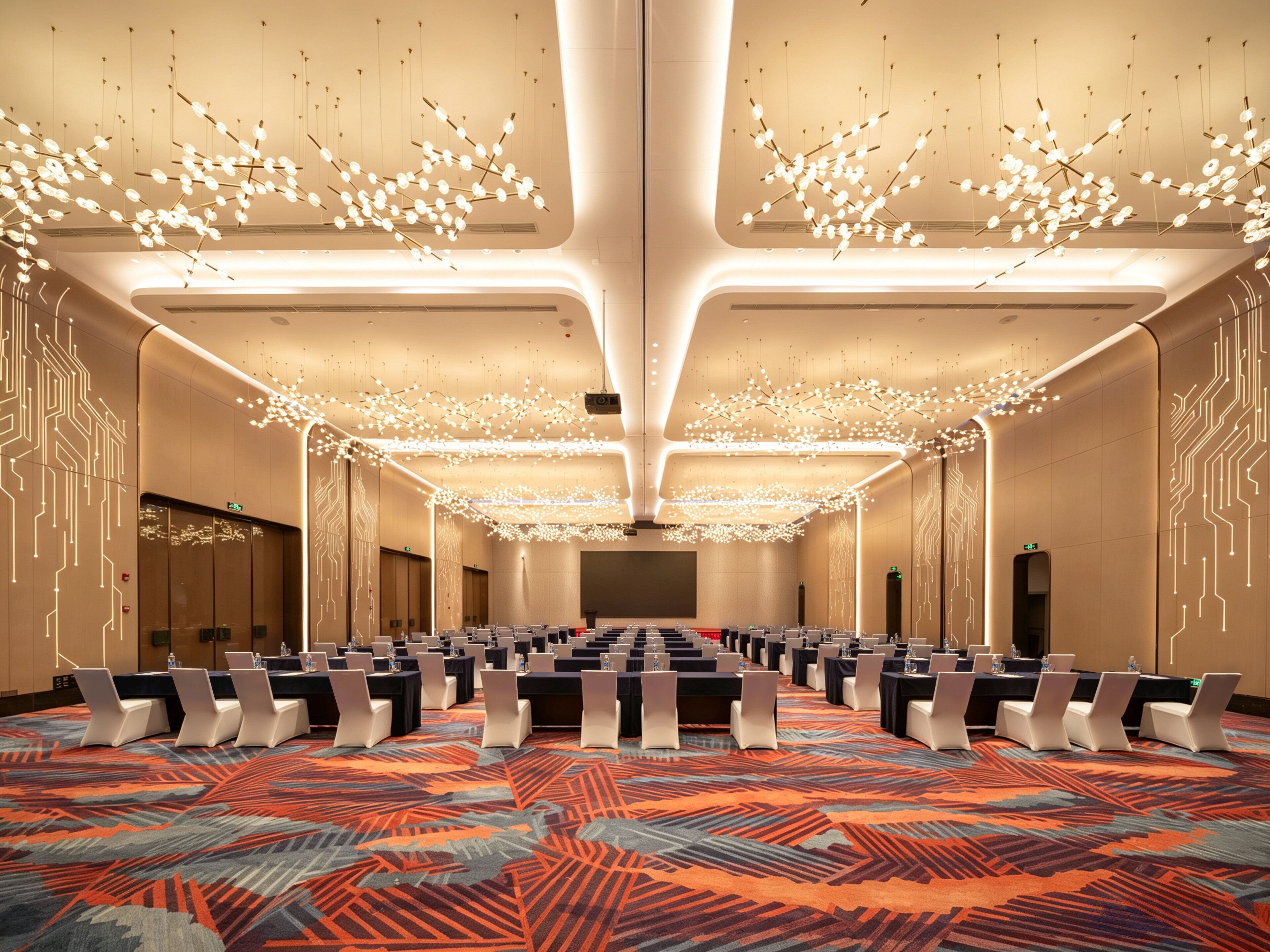 Une salle de conférence avec des rangées de tables et de chaises, une moquette à motifs et un lustre moderne.
