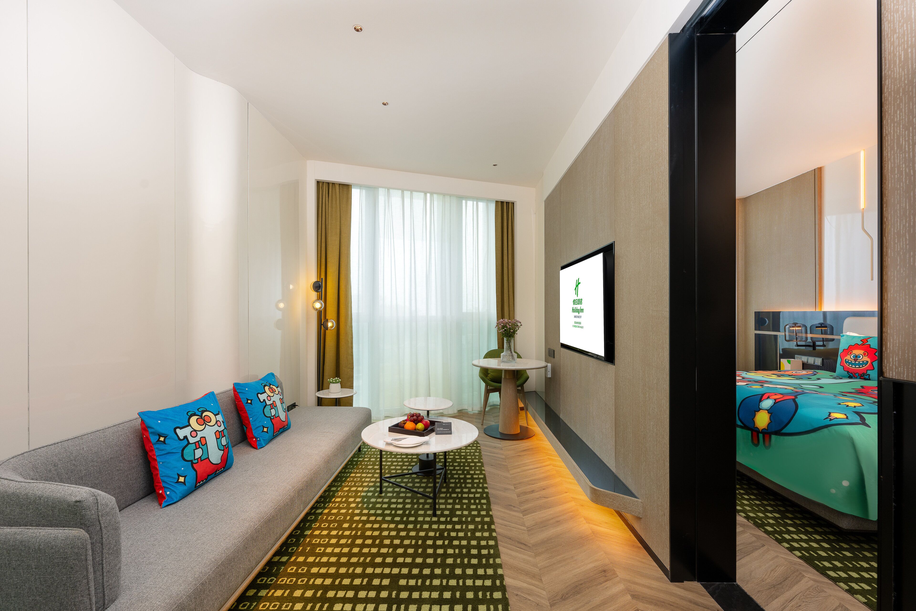 Una habitación de hotel moderna con un sofá gris, almohadas coloridas, una pequeña mesa blanca con fruta, una alfombra verde estampada y una cama con colcha azul.