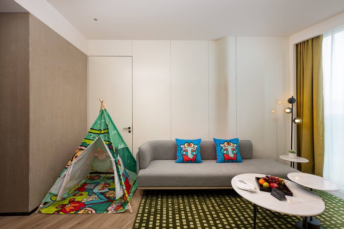 A modern hotel room with a grey sofa, two blue pillows, a small round table with a fruit bowl, a green patterned rug, and a tent-like play area.