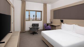Habitación de hotel con cama, escritorio, silla, televisor y ventana con vistas a edificios y palmeras.