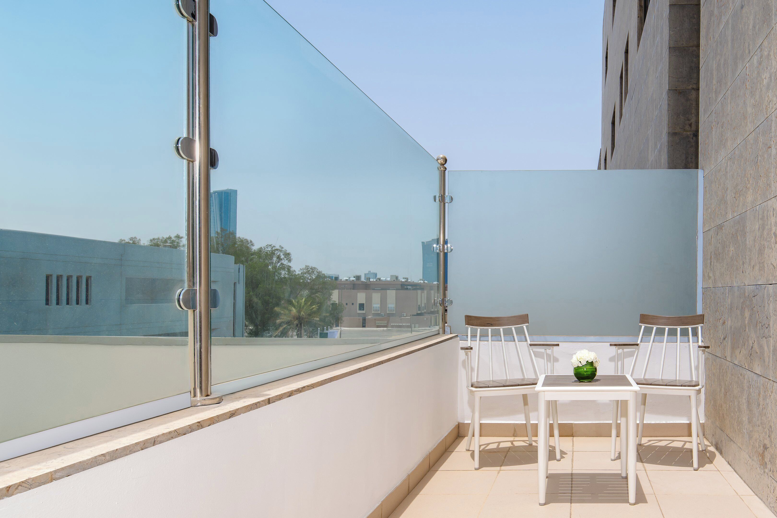 Un balcon avec deux chaises blanches et une petite table, une paroi en verre et une vue sur des immeubles et des arbres.