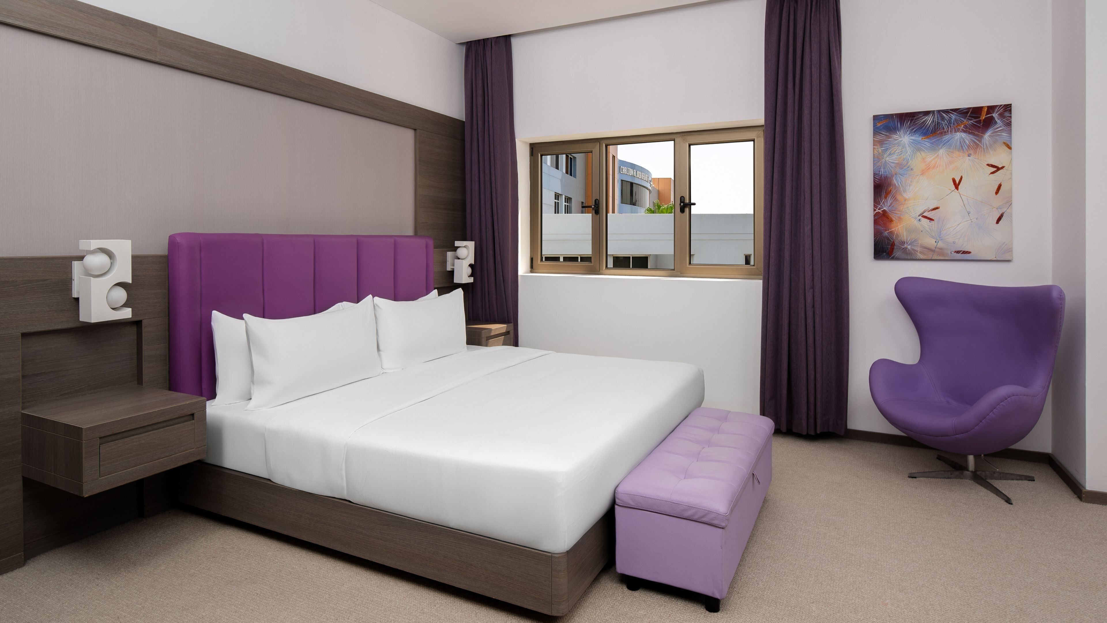 Et moderne hotelværelse med en stor seng, et lille sengebord, en lilla stol og et vindue med udsigt til bygninger.