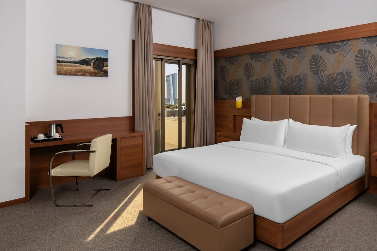 Ein Hotelzimmer mit einem großen Bett, einem Schreibtisch mit Stuhl, einem kleinen Tisch, einer Bank und Blick auf die Stadt durch eine Glastür.