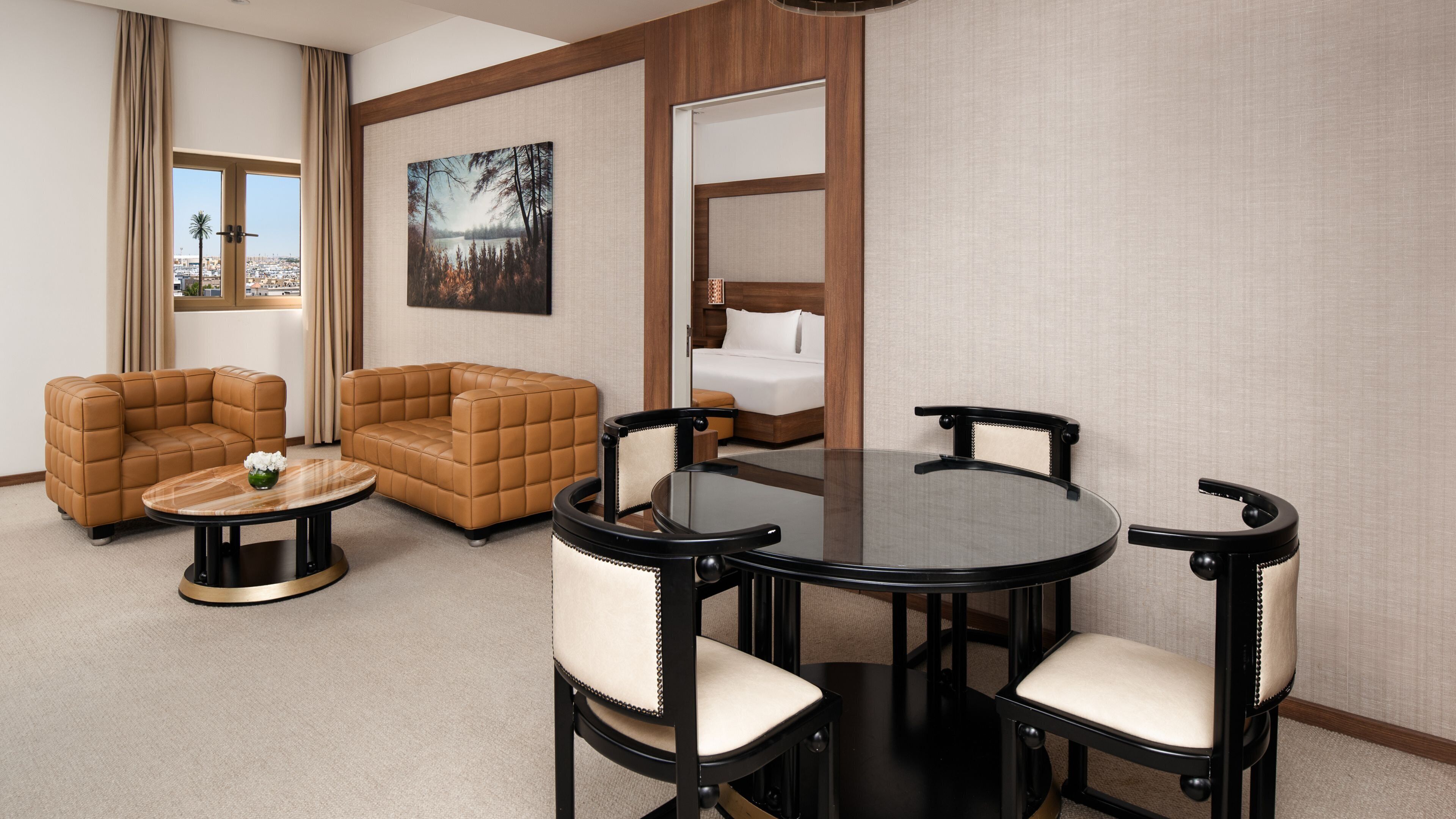 Une chambre d’hôtel avec un canapé en cuir marron, une table de salle à manger ronde entourée de quatre chaises, un lit avec du linge de maison blanc et une grande fenêtre avec des rideaux.