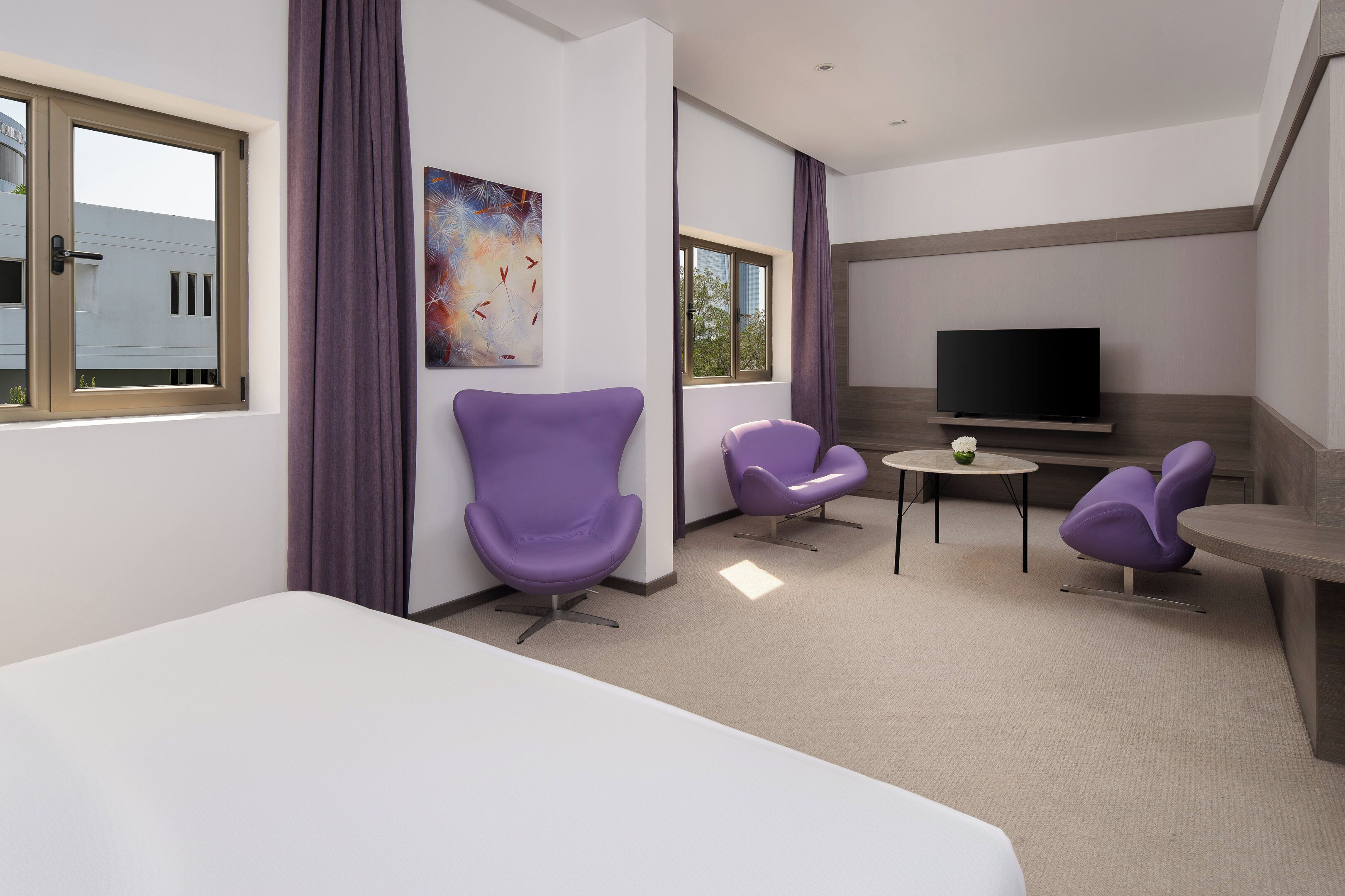 Een moderne hotelkamer met een groot bed, twee paarse stoelen, een kleine tafel, een flatscreen televisie en een schilderij aan de muur.