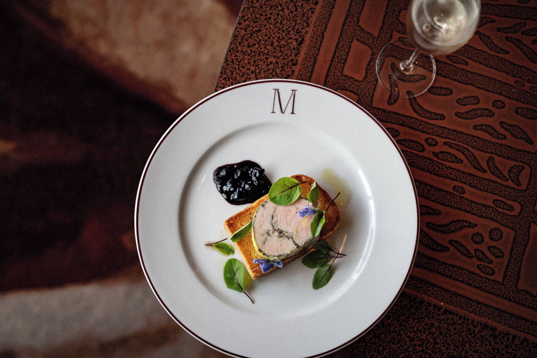 A plate with a dish, a glass of white wine, and a patterned tablecloth.