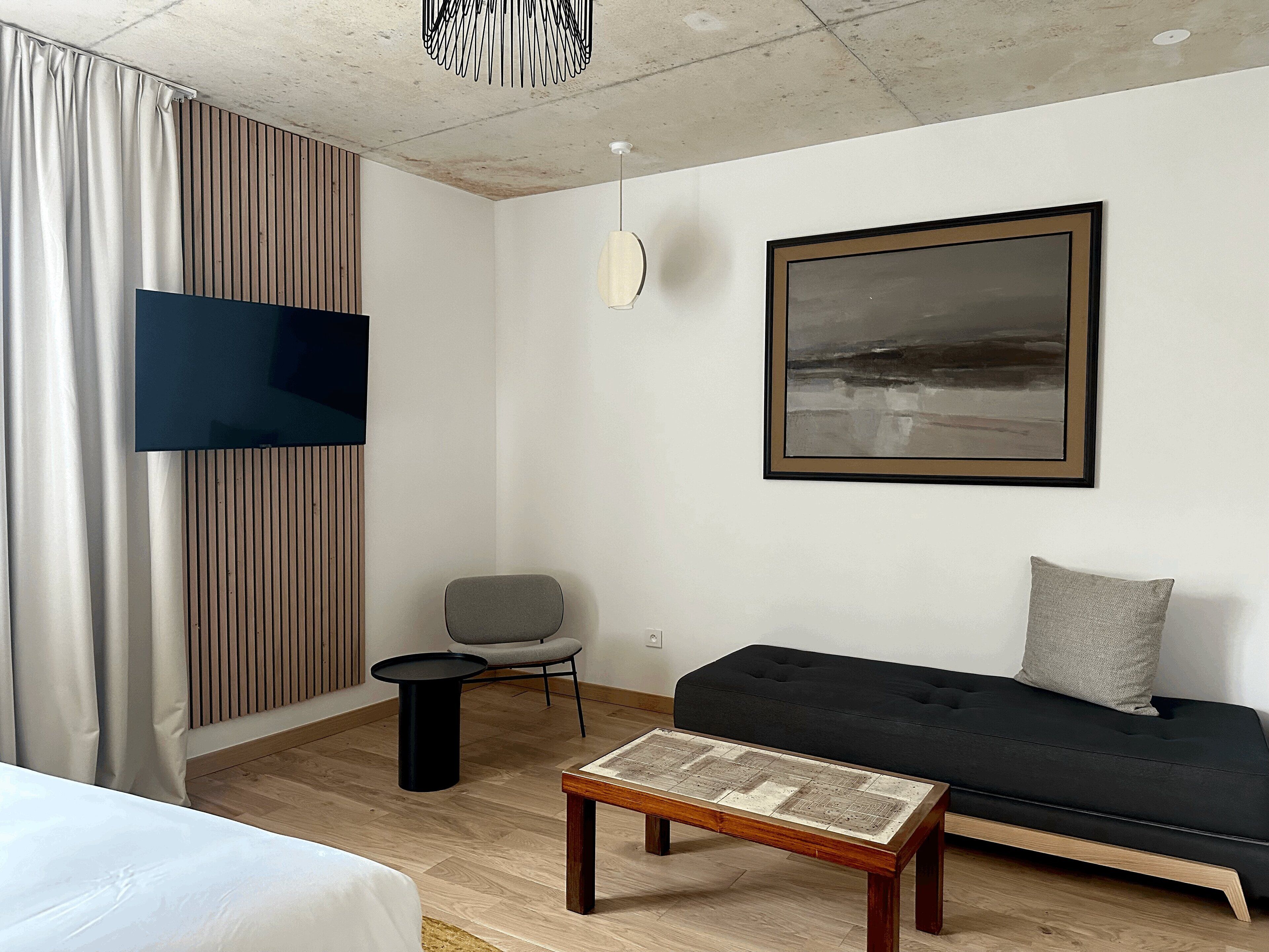 Een moderne hotelkamer met een houten vloer, een zwarte bank, een grijze fauteuil, een klein rond tafeltje en een groot abstract schilderij aan de muur.