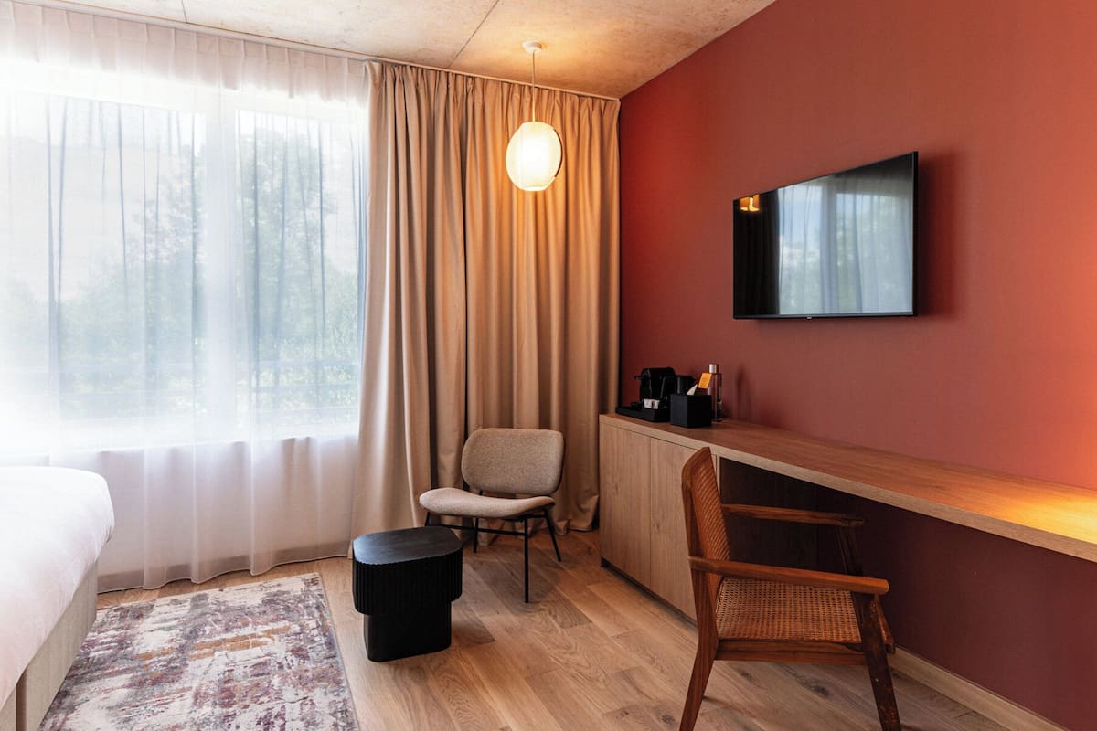 A hotel room with a bed, a desk with a chair, a television, and a window with curtains.
