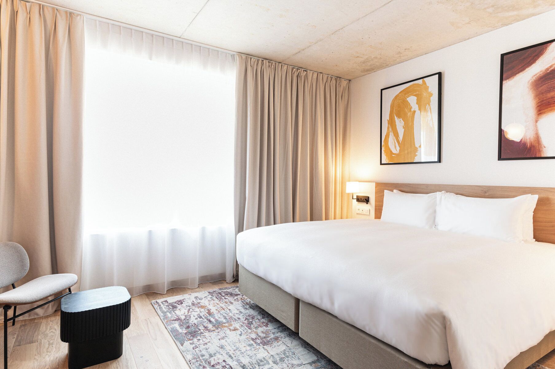 Habitación de hotel con una cama grande, una silla, una mesita y vistas al exterior a través de cortinas translúcidas.