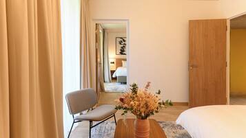 Una habitación de hotel con cama, una silla, una mesa de madera con un florero y una puerta que da a otra habitación.