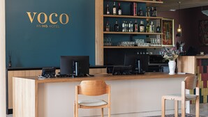 A bar area with a wooden counter, two chairs, a laptop, and a wall-mounted shelf with bottles and glasses.