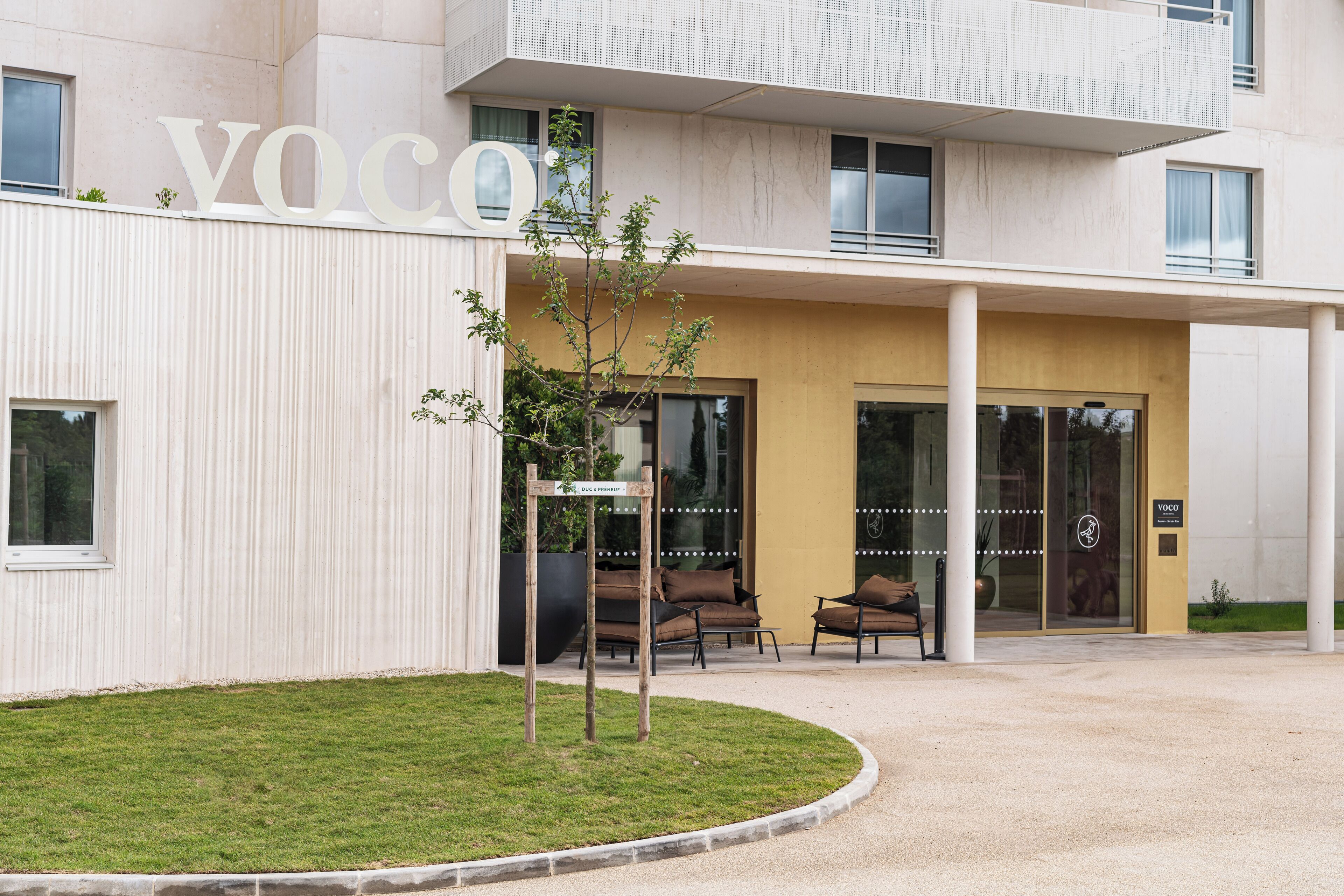 A modern building with a prominent 'VOCO' sign, a well-maintained lawn, and a seating area with chairs and a table.
