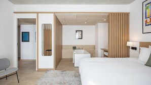 A modern hotel room with a large bed, a small desk, a chair, a bathroom with a bathtub, and a wooden panel wall.