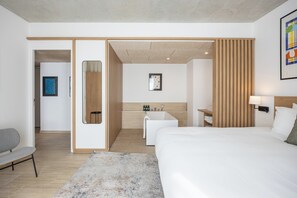 A modern hotel room with a large bed, a small desk, a chair, a bathroom with a bathtub, and a wooden panel wall.