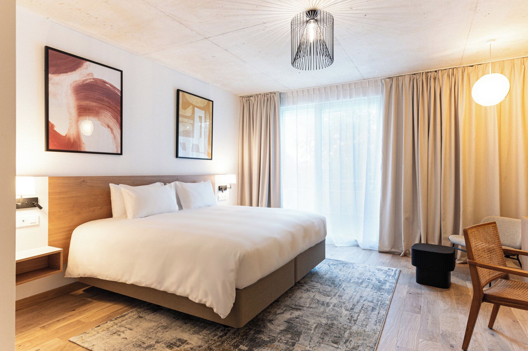 A hotel room with a large bed, a wooden headboard, a bedside table, a chair, and a large window with curtains.