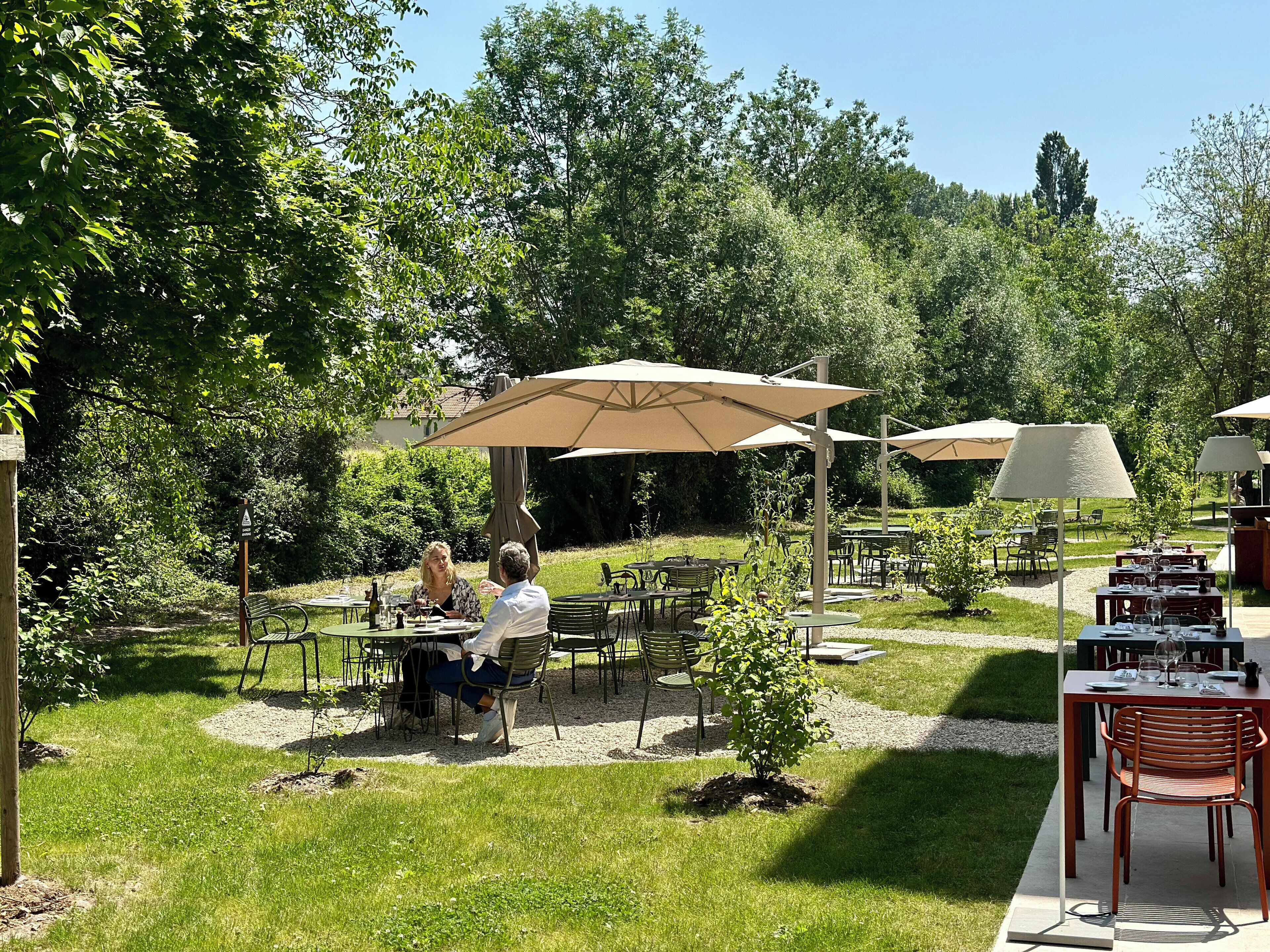 An outdoor dining area with tables and chairs, surrounded by greenery and trees.