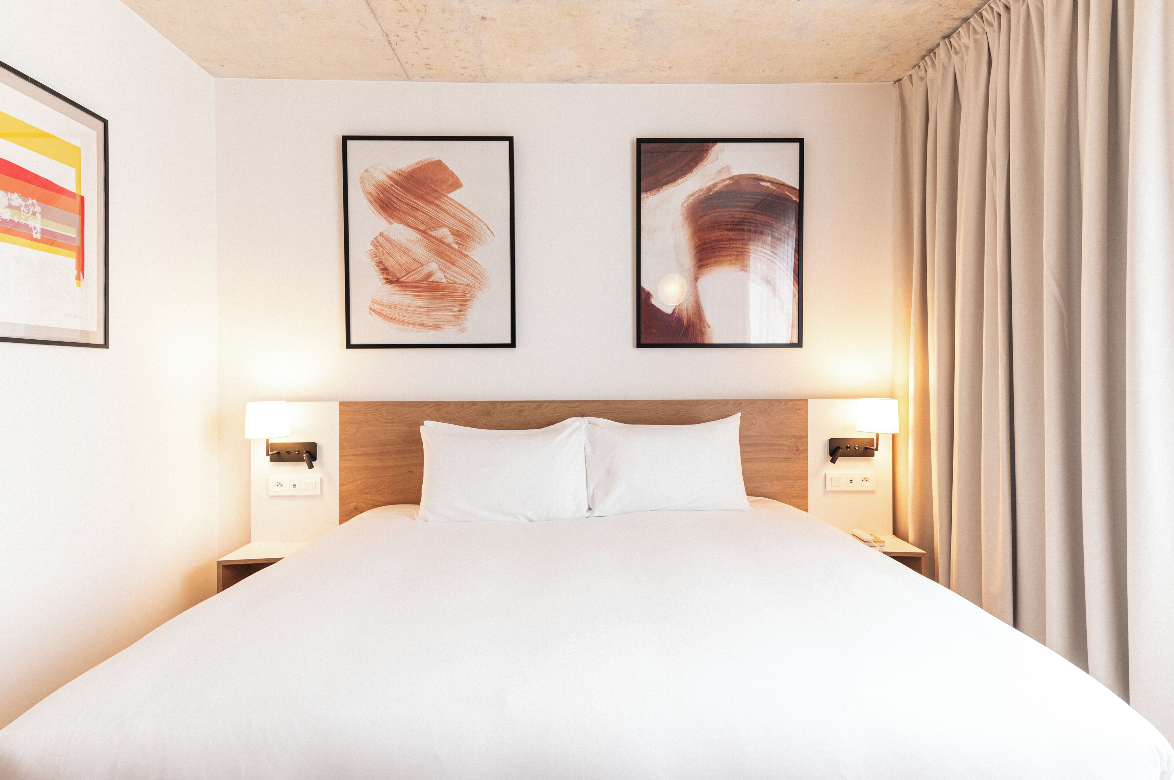 A hotel room with a large bed, two framed artworks on the wall, and a nightstand with a lamp.