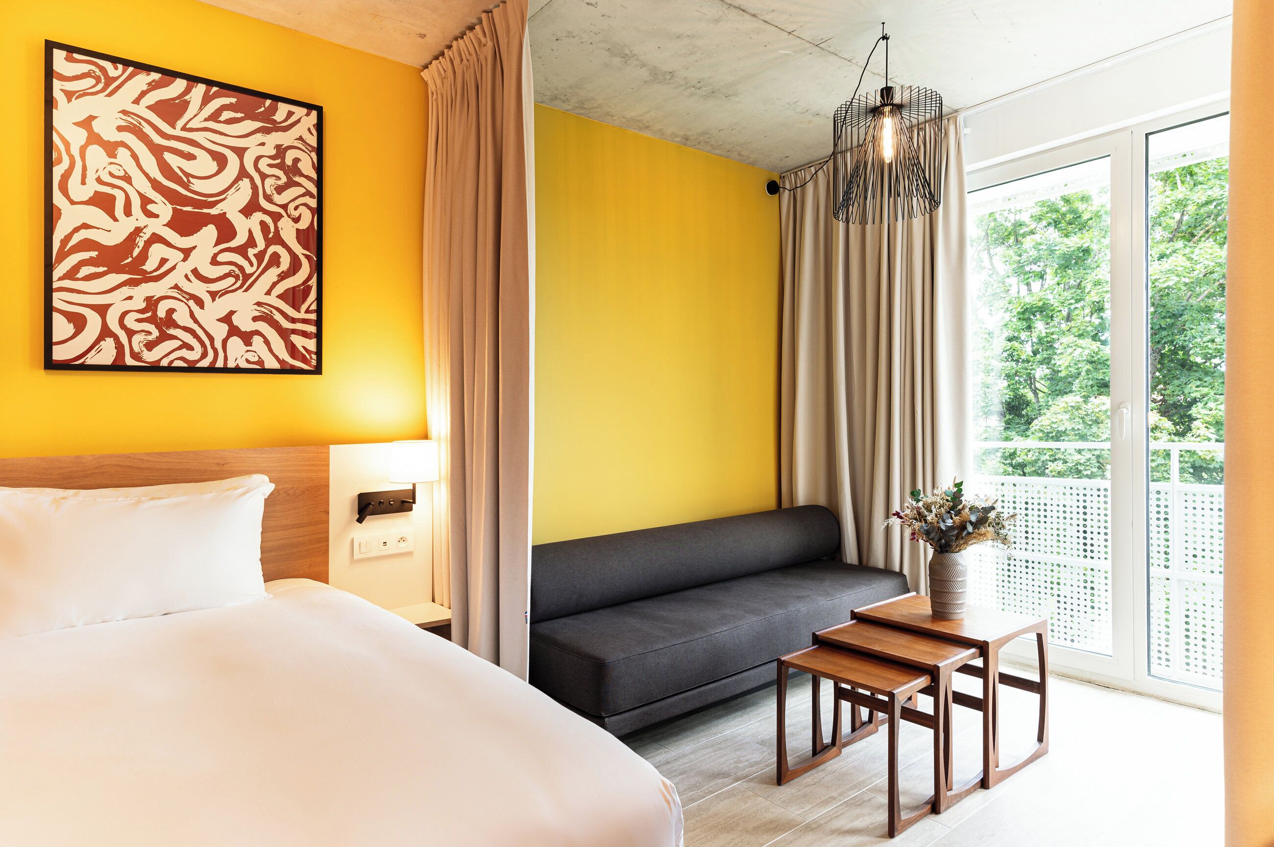A hotel room with a bed, a sofa, a small table with a vase, and a view of greenery through the window.
