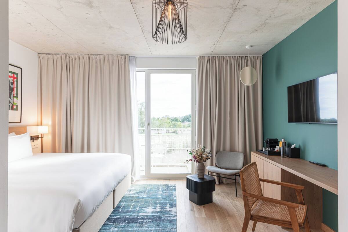 A hotel room with a bed, a desk, a chair, a small table with a vase, and a large window with curtains.