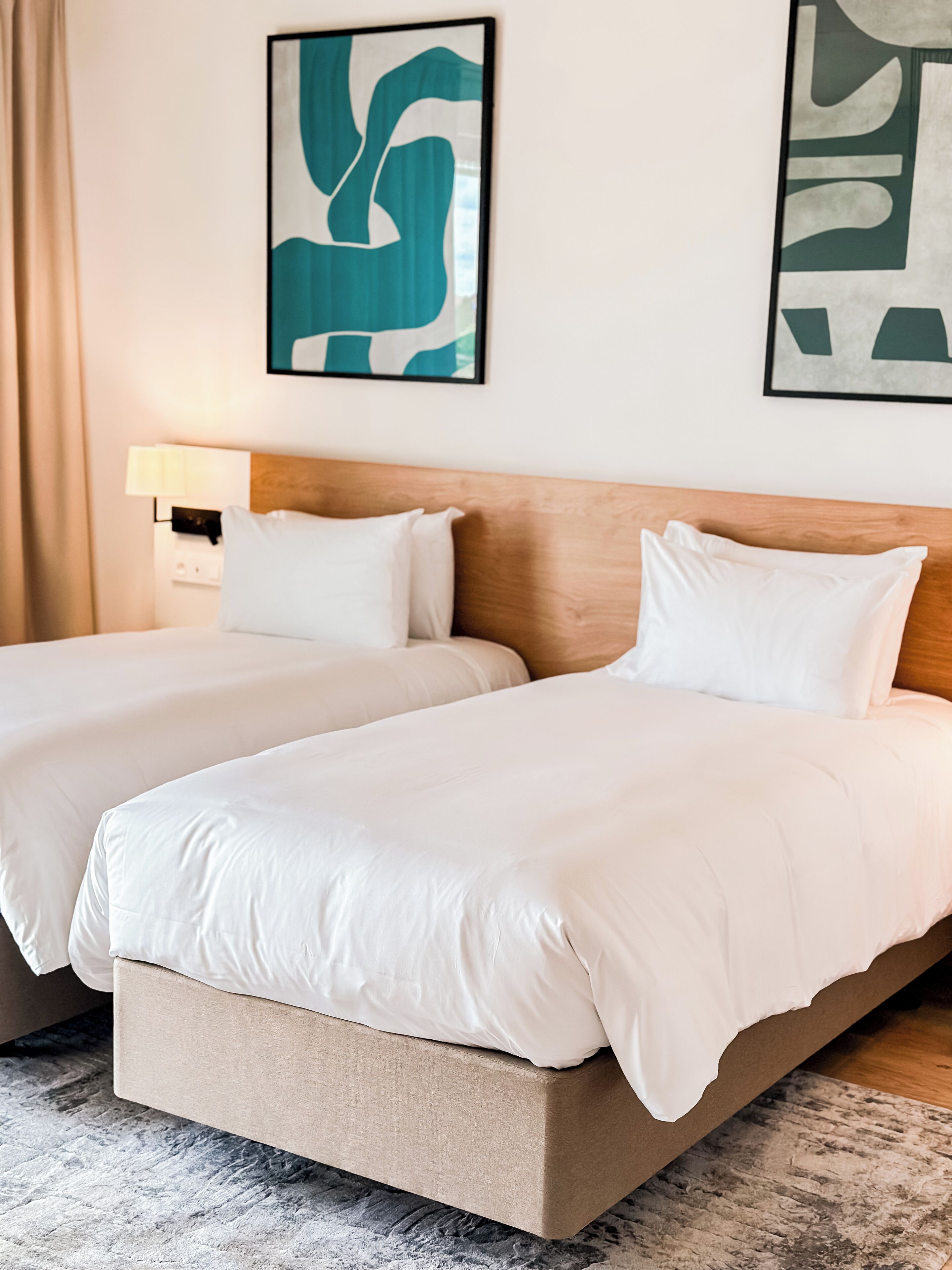 A hotel room with two beds, a wooden headboard, a nightstand with a lamp, and abstract wall art.