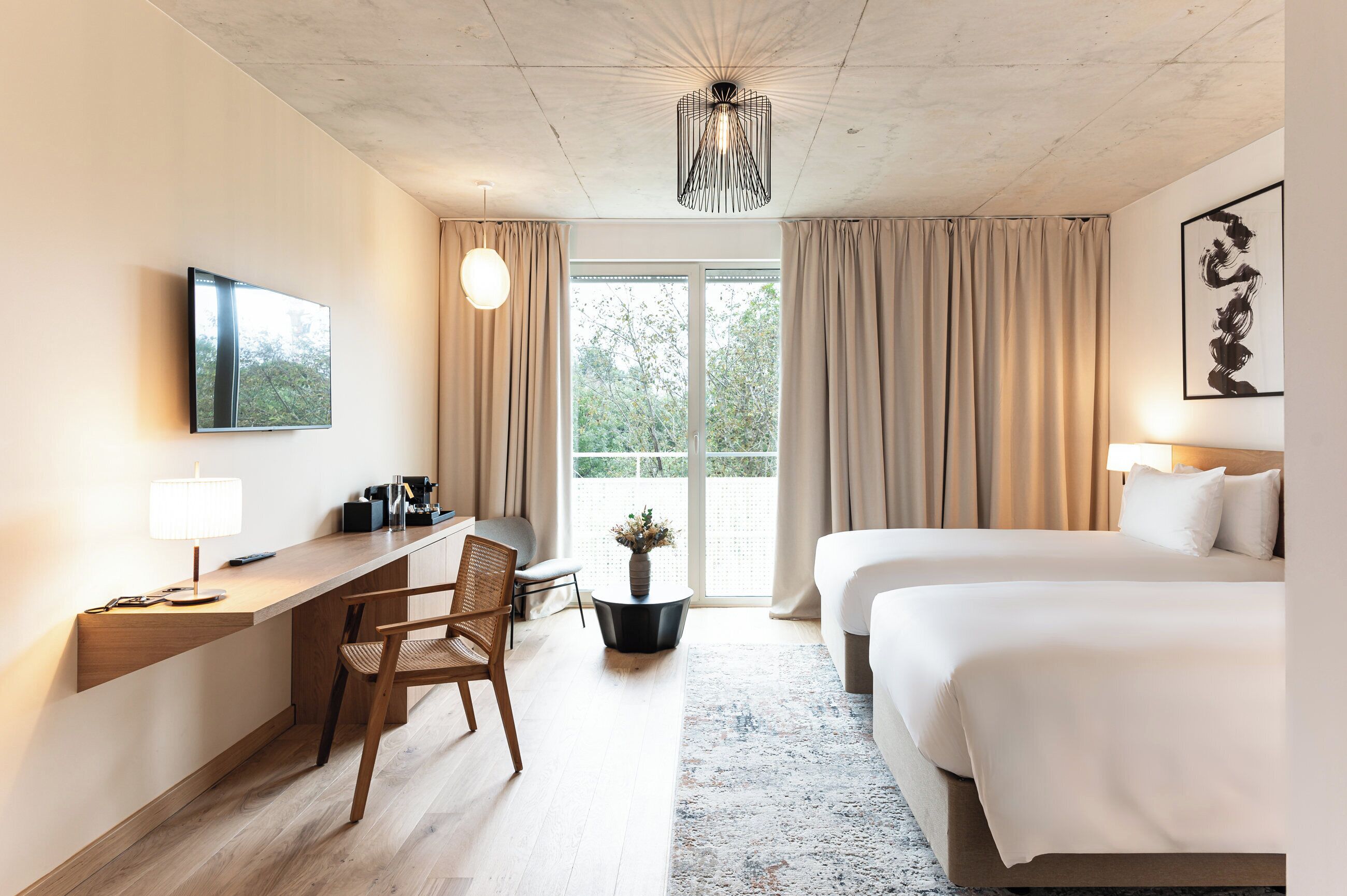 Una habitación de hotel con escritorio, dos camas, televisor, candelabro y vistas por la ventana.