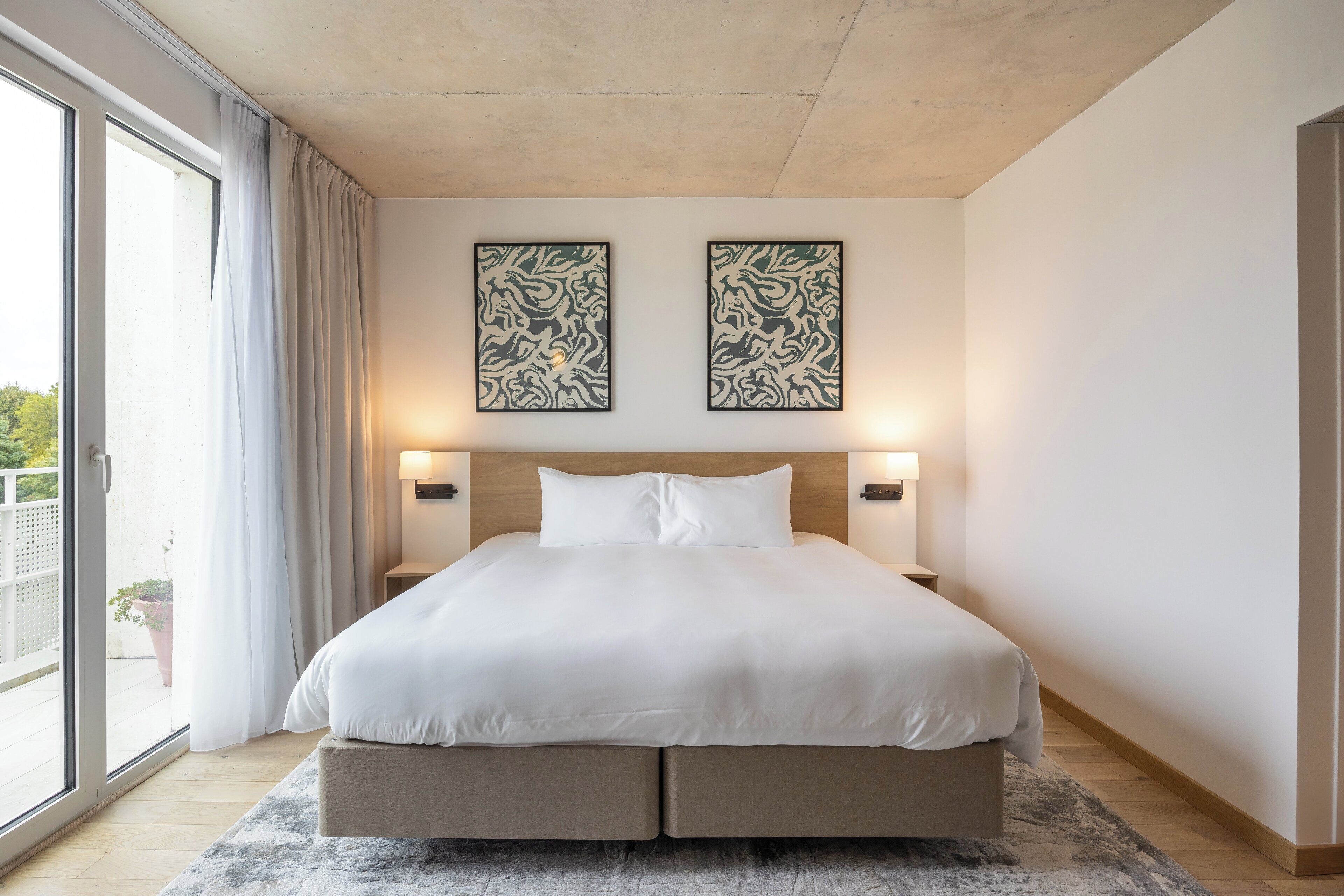 Une chambre moderne avec un grand lit, deux lampes fixées au mur et une porte-fenêtre coulissante donnant sur un balcon.