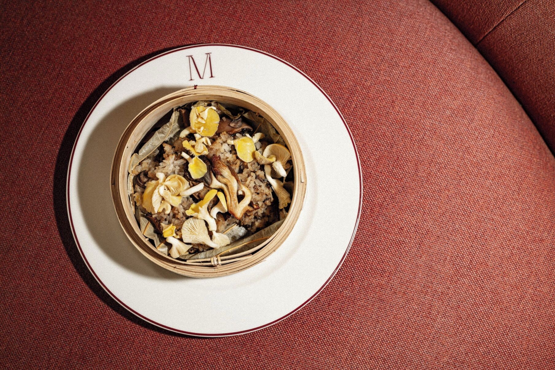A bowl of food with mushrooms and lemon slices on a plate with a letter 'M' on it, placed on a textured surface.
