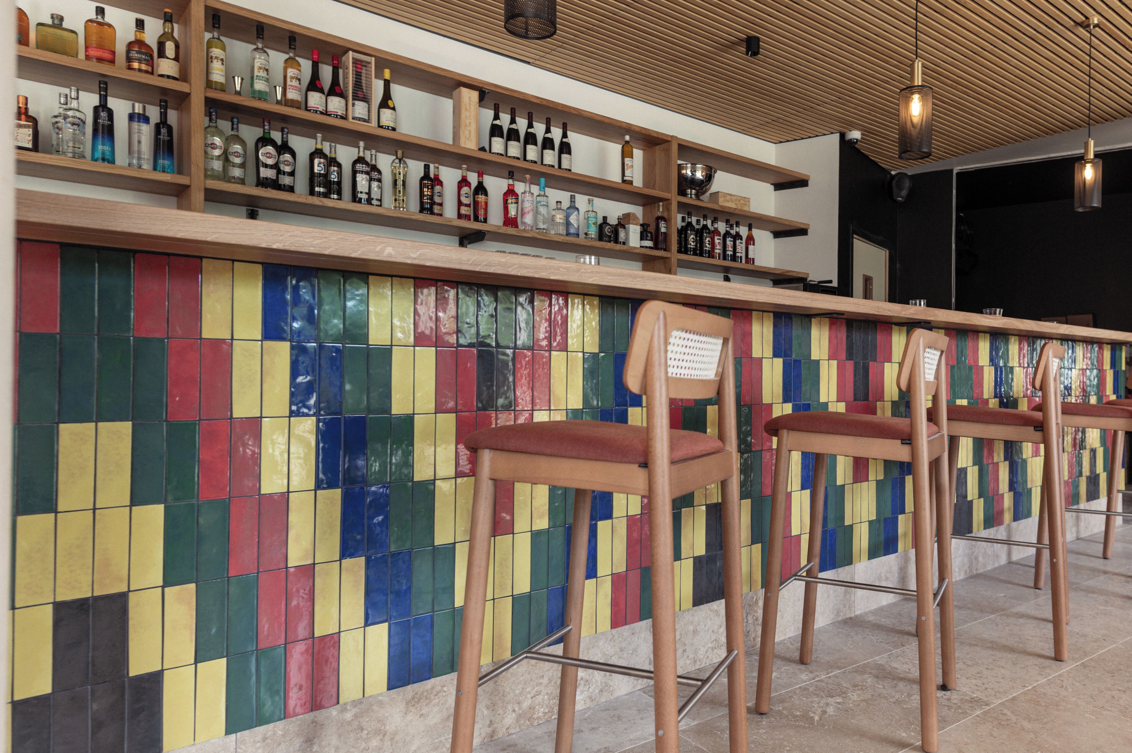 A bar area with wooden shelves displaying various bottles, colorful tiled wall, and bar stools.