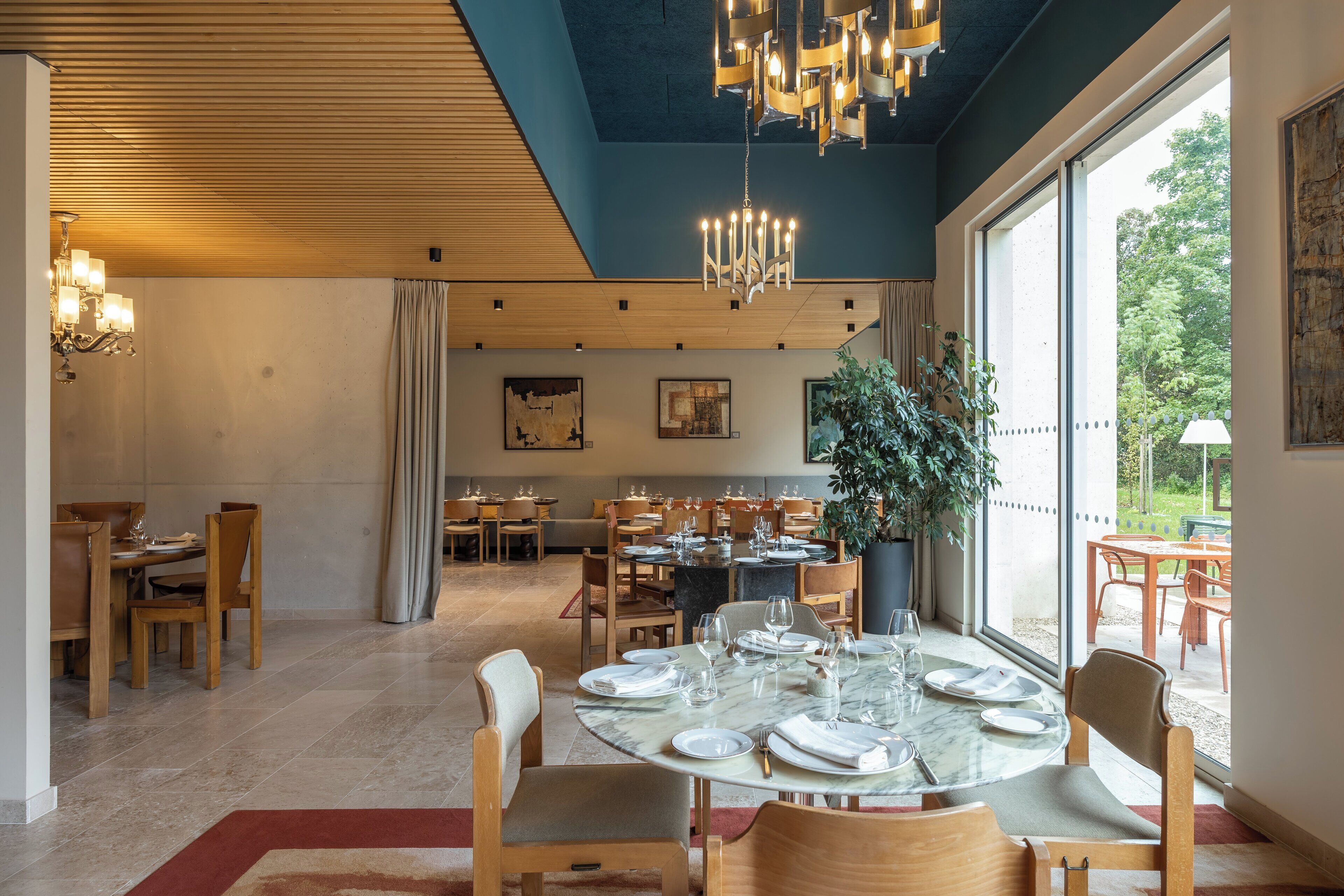 Un comedor espacioso con mesas redondas dispuestas para comer, una planta grande en maceta y vistas al exterior a través de amplios ventanales.