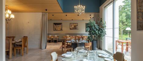 Un comedor espacioso con mesas redondas dispuestas para comer, una planta grande en maceta y vistas al exterior a través de amplios ventanales.