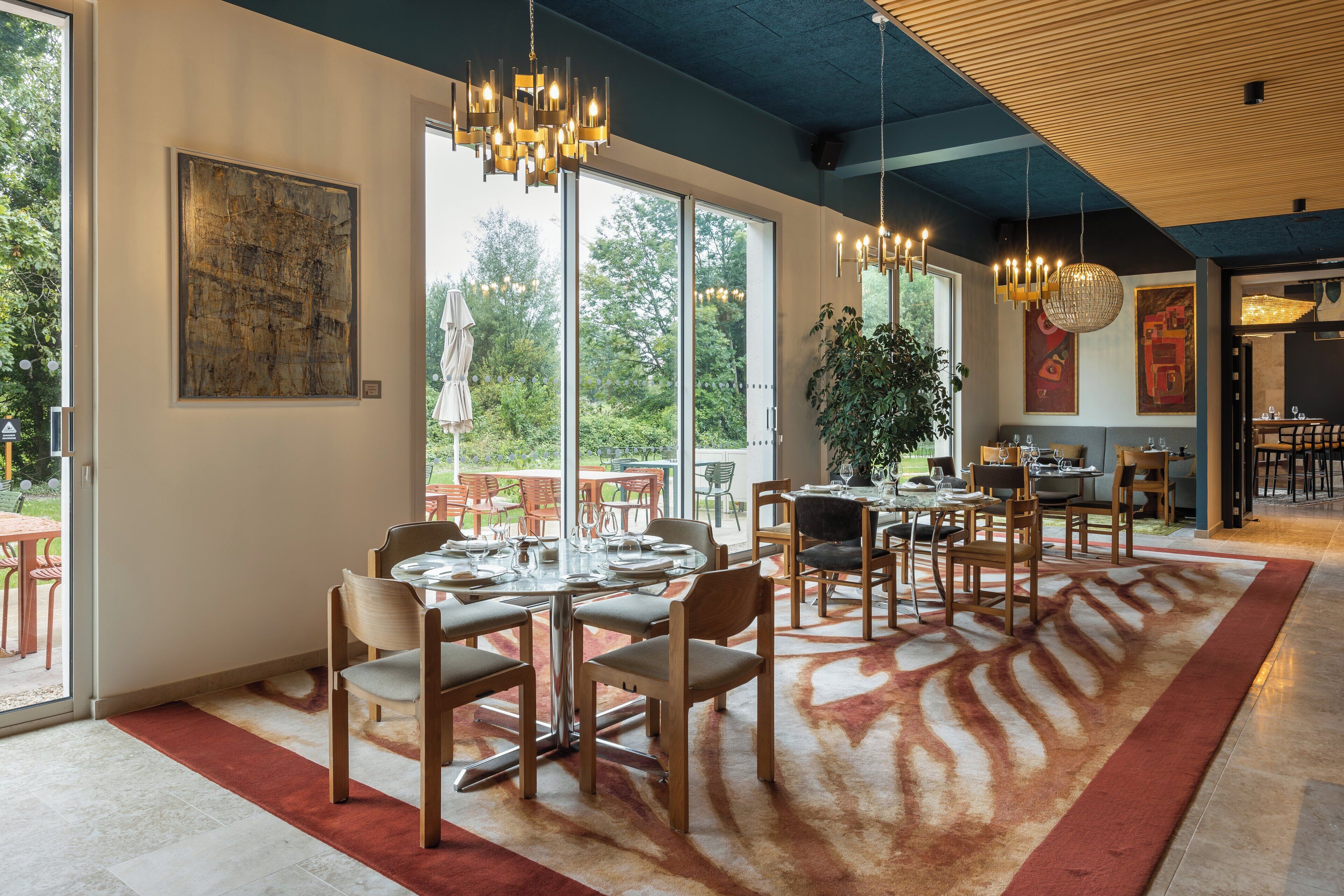 A spacious dining area with multiple tables and chairs, large windows offering a view of the outdoors, and a modern chandelier.