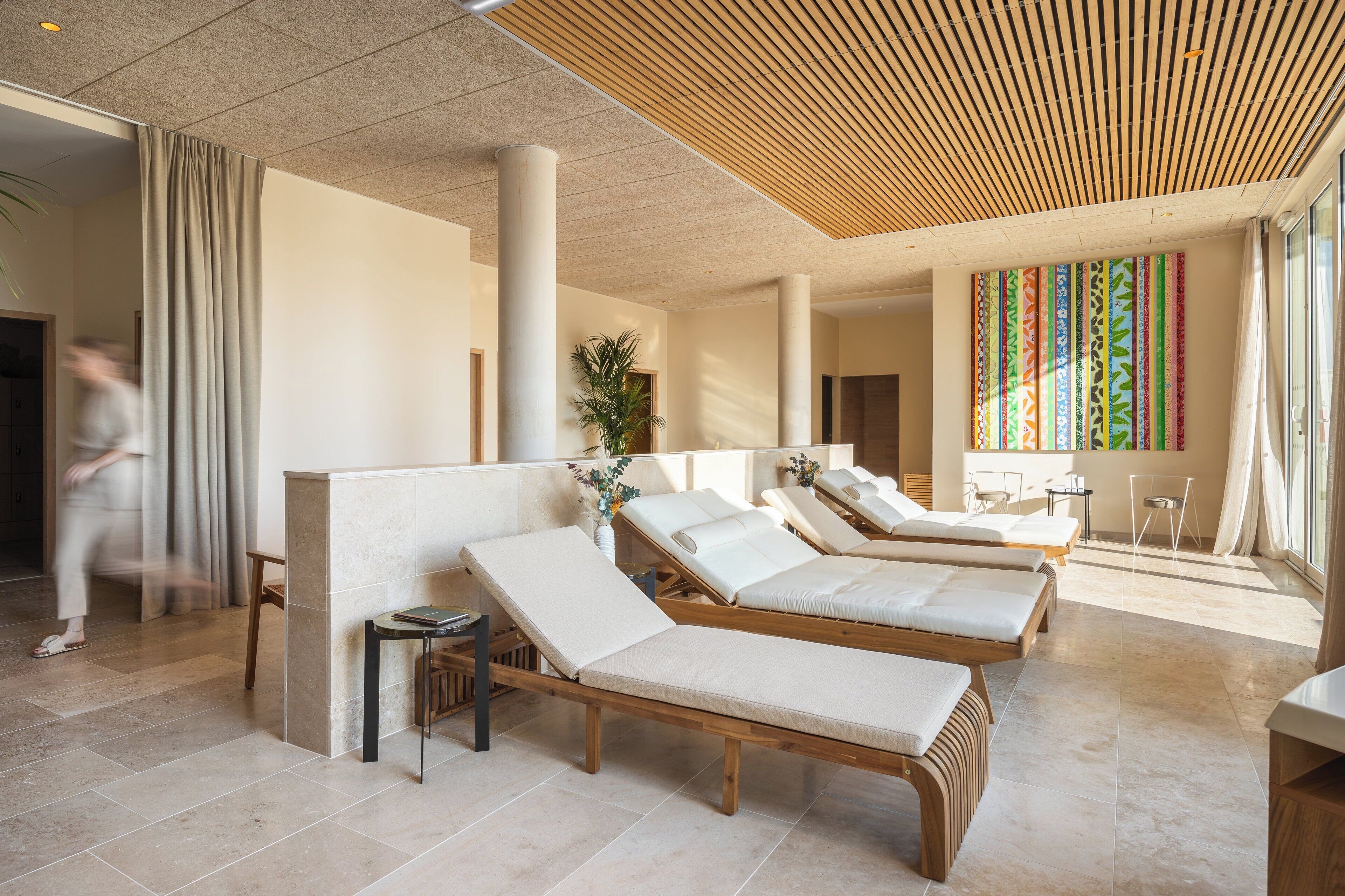 Un espace détente avec des chaises longues en bois, une grande plante et une œuvre d’art murale aux couleurs vives.