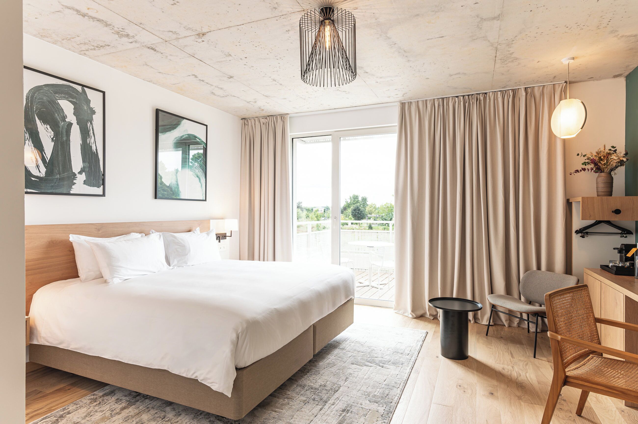 A modern hotel room with a large bed, a desk, a chair, and a view of the outdoors.