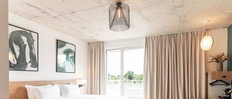 A modern hotel room with a large bed, a desk, a chair, and a view of the outdoors.