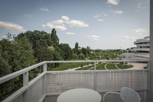 Un balcon avec une table et une chaise blanches, donnant sur un parc arboré et sur lequel on aperçoit, au loin, un bâtiment moderne.