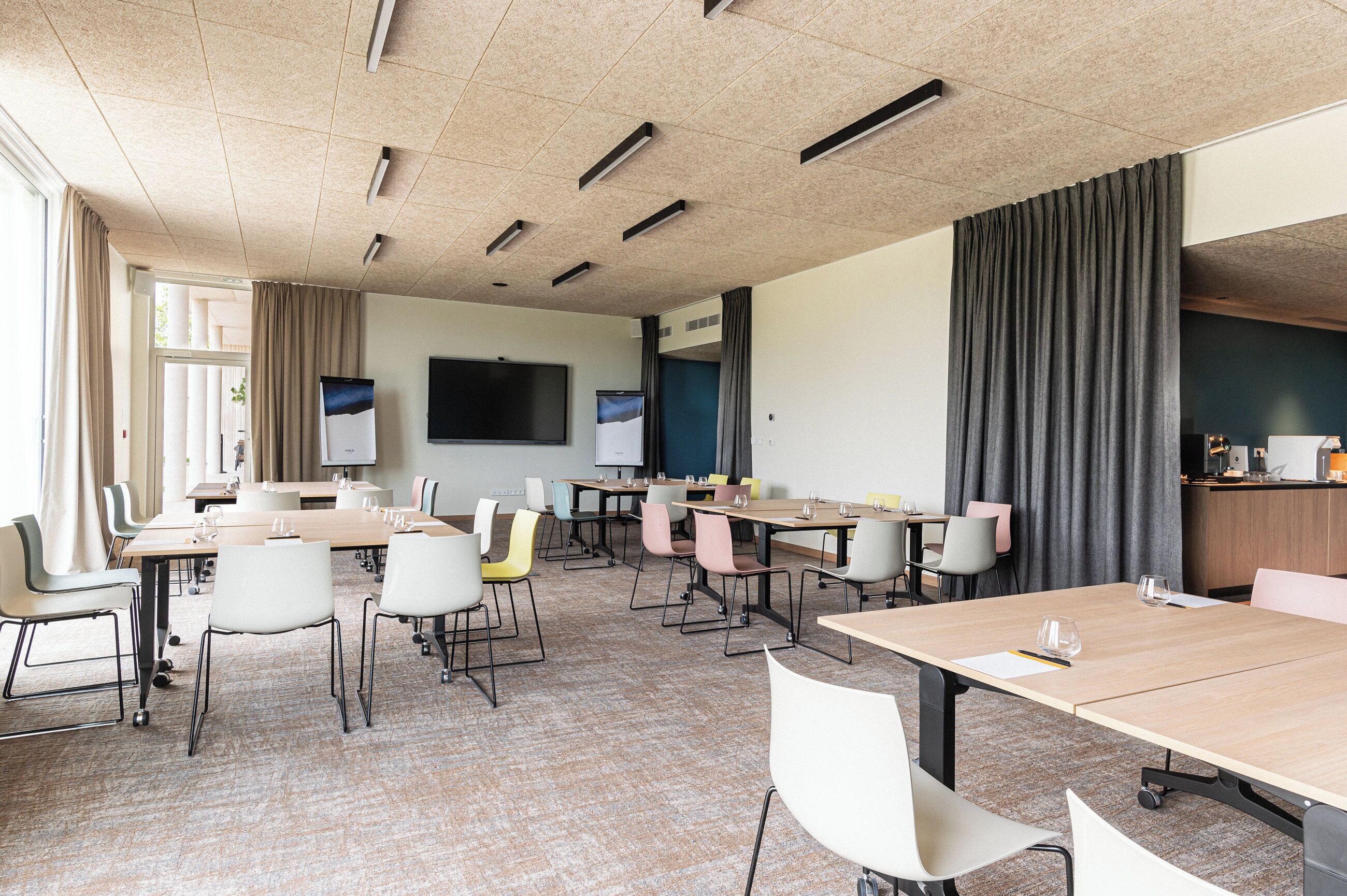 Une salle de conférence équipée de plusieurs tables et chaises, d’un grand téléviseur à écran plat et offrant une vue sur l’extérieur grâce à de grandes fenêtres.