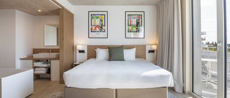 A modern hotel room with a large bed, a built-in desk, a bathroom, and large windows offering a city view.